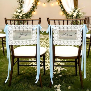 Op de afbeelding: Twee houten stoelen met witte kussens zijn versierd voor een bruiloft. Elke stoel heeft een bord met de woorden "Just" en "Married" in elegant schrift, omlijst door wit kant en vastgebonden met lichtblauwe linten.