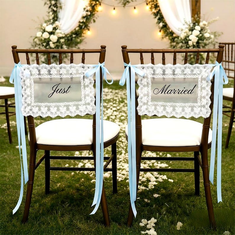 Op de afbeelding: Twee houten stoelen met witte kussens zijn versierd voor een bruiloft. Elke stoel heeft een bord met de woorden "Just" en "Married" in elegant schrift, omlijst door wit kant en vastgebonden met lichtblauwe linten.