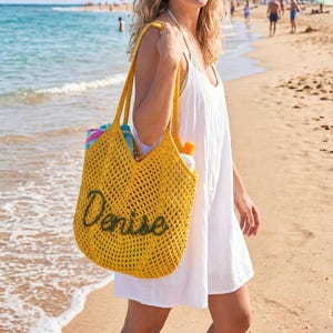 May include: A yellow crochet tote bag with the name "Denise" in green script. The bag is filled with beach essentials and is being carried on a sandy beach near the ocean. The bag has long handles.