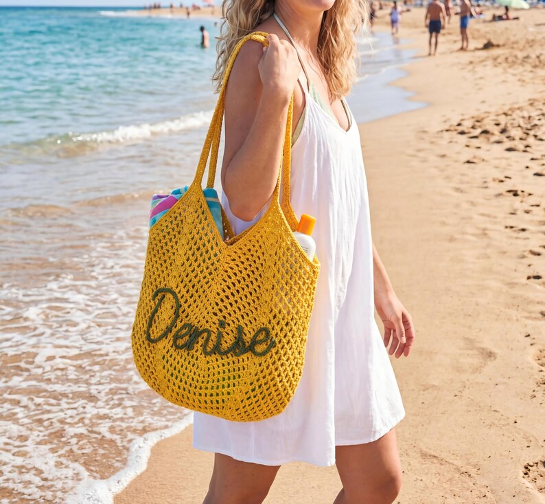 May include: A yellow crochet tote bag with the name "Denise" in green script. The bag is filled with beach essentials and is being carried on a sandy beach near the ocean. The bag has long handles.