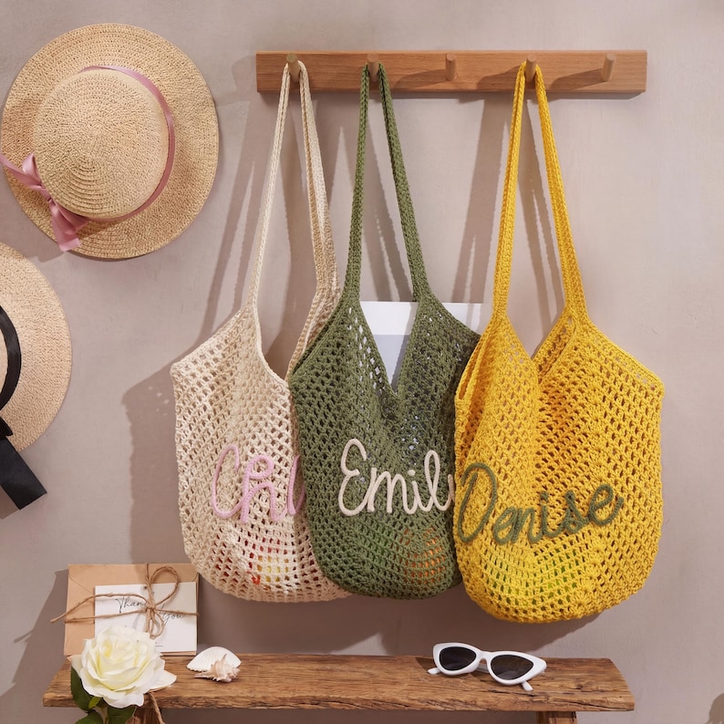 May include: Three crocheted tote bags in cream, olive green, and yellow hang on a wooden rack. Each bag has a name embroidered on it in a contrasting color. Two straw hats with pink ribbons are visible in the background.
