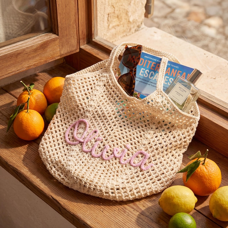 May include: A beige crocheted tote bag with the name "Olivia" in pink script. The bag contains sunglasses, a book, and a perfume bottle. Oranges, lemons, and limes are arranged around the bag on a wooden surface.