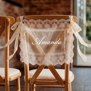 Op de afbeelding: Twee houten stoelen met cr&egrave;mekleurige kussens. De rugleuningen zijn versierd met transparante, cr&egrave;mekleurige stoffen panelen afgezet met kant en vastgebonden met cr&egrave;mekleurige linten. De naam "Amanda" staat op de stof.