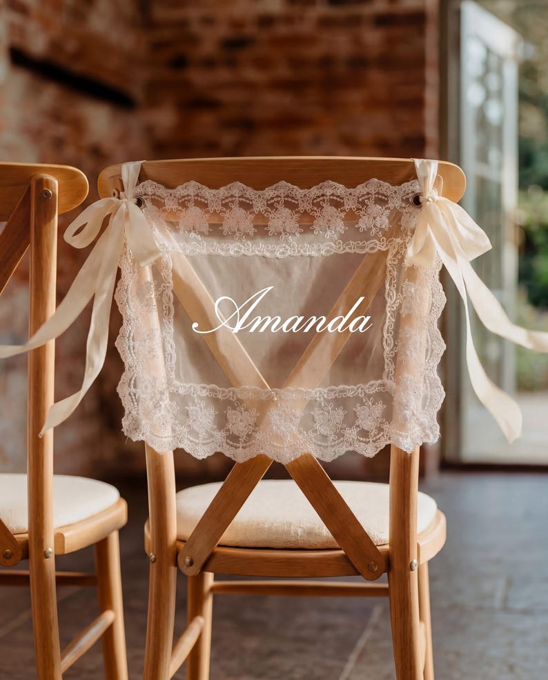 Op de afbeelding: Twee houten stoelen met cr&egrave;mekleurige kussens. De rugleuningen zijn versierd met transparante, cr&egrave;mekleurige stoffen panelen afgezet met kant en vastgebonden met cr&egrave;mekleurige linten. De naam "Amanda" staat op de stof.