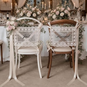 Op de afbeelding: Twee stoelen versierd voor een bruiloft. E&eacute;n witte stoel heeft een bord met "Just" en de andere bruine stoel heeft een bord met "Married". Beide borden zijn omlijst met kant en linten. Bloemschikkingen en kaarsen staan op de achtergrond.