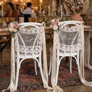 Op de afbeelding: Twee witte stoelen met kanten rugleuningen en cr&egrave;mekleurige linten zijn versierd voor een bruiloft. Op de rugleuningen van de stoelen staan de woorden "Just" en "Married". Bloemsierkunst van roze en witte rozen siert de stoelen. De stoelen staan aan een tafel met kaarsen en glaswerk.