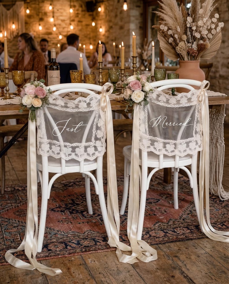 Op de afbeelding: Twee witte stoelen met kanten rugleuningen en cr&egrave;mekleurige linten zijn versierd voor een bruiloft. Op de rugleuningen van de stoelen staan de woorden "Just" en "Married". Bloemsierkunst van roze en witte rozen siert de stoelen. De stoelen staan aan een tafel met kaarsen en glaswerk.