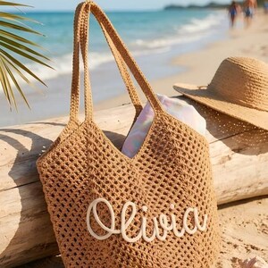 May include: A tan crochet tote bag with the name "Olivia" in white script. The bag has long handles and is sitting on a sandy beach next to a straw hat. The background shows the ocean and a palm frond.