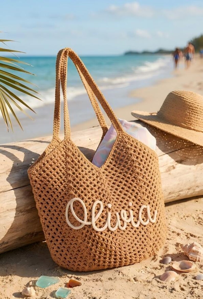 May include: A tan crochet tote bag with the name "Olivia" in white script. The bag has long handles and is sitting on a sandy beach next to a straw hat. The background shows the ocean and a palm frond.