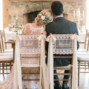 Op de afbeelding: Trouwreceptie met een bruid en bruidegom aan een tafel. De rugleuningen van hun stoelen zijn versierd met kanten panelen met de tekst "Just" en "Married". Cr&egrave;mekleurige linten hangen van de stoelen.