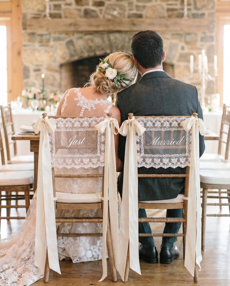 Op de afbeelding: Trouwreceptie met een bruid en bruidegom aan een tafel. De rugleuningen van hun stoelen zijn versierd met kanten panelen met de tekst "Just" en "Married". Cr&egrave;mekleurige linten hangen van de stoelen.