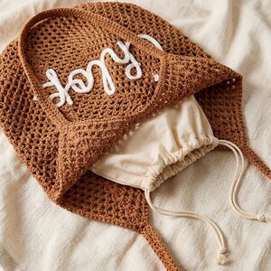 May include: A brown crocheted tote bag with the word "Joy" embroidered in white. The bag has long handles and a drawstring pouch inside. The bag is made of a woven material and is sitting on a cream-colored surface.