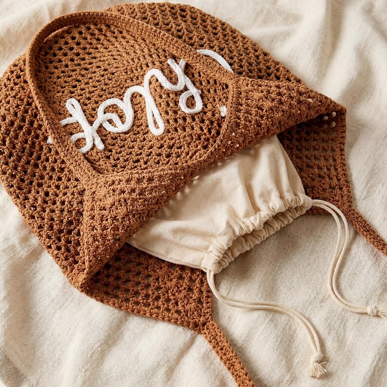 May include: A brown crocheted tote bag with the word "Joy" embroidered in white. The bag has long handles and a drawstring pouch inside. The bag is made of a woven material and is sitting on a cream-colored surface.