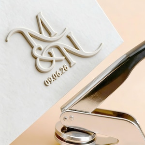 May include: A cream-coloured embossed design featuring the intertwined letters "M" and "N" and the date "09.06.26". A shiny silver and black embosser tool is in the foreground, suggesting a wedding or event stationery theme.