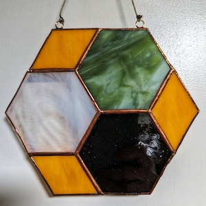 May include: A hexagonal stained glass window hanging with copper framing. The window is made of six pieces of glass: two yellow, one white, one green, and two black.