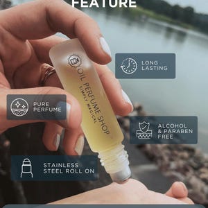 K&ouml;nnte beinhalten: Nahaufnahme einer Hand, die eine kleine Glasflasche mit einem Roll-on-Applikator h&auml;lt. Die Flasche ist mit "Oil Perfume Shop Simply Magical" beschriftet. Die Flasche ist von Symbolen umgeben, die die Eigenschaften des Produkts hervorheben, darunter "Reines Parfum", "Lang anhaltend", "Roll-on aus Edelstahl" und "Alkohol- und parabenfrei".