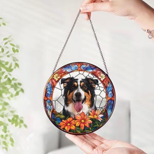 May include: A round stained glass window featuring a dog with black, brown, and white fur, a pink tongue, and a floral border. The window is held by a chain and has a colorful design with blue, orange, and yellow flowers.
