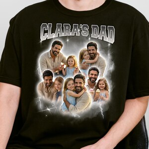 May include: Black t-shirt with the text "CLARA'S DAD" in white and gray. The shirt features a collage of photos of a man and two young girls, surrounded by a cloudy, lightning-bolt design.