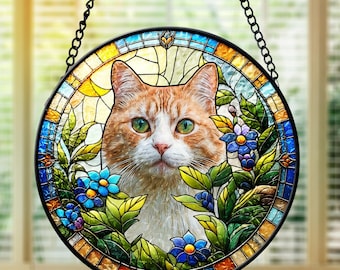May include: Round stained glass window featuring an orange and white cat with green eyes, surrounded by blue flowers and green leaves. The window is framed in black and hangs from a black chain.