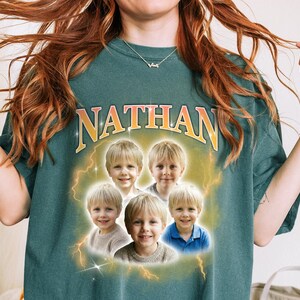 May include: A teal-colored t-shirt with the name "NATHAN" in gold lettering. The shirt features a graphic of five children's portraits surrounded by lightning bolts. The children are wearing various colored shirts.