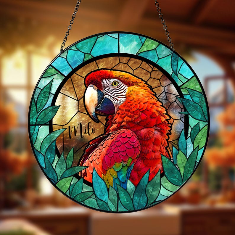 May include: A round stained glass window featuring a vibrant macaw parrot design. The parrot has red, orange, and yellow feathers, with a white beak and a green eye. The name "Milo" is written on the glass. The window is framed with green leaves and a blue border.