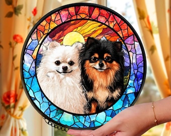 May include: A round, stained-glass window featuring two Pomeranian dogs. One dog is white, the other is black and brown. The background depicts a sunset with vibrant colors of red, orange, yellow, and blue.