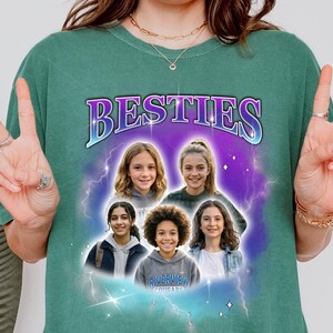 May include: Green t-shirt with the word "BESTIES" in a purple and blue design. The shirt features a graphic of five smiling girls in front of a purple and blue background with lightning bolts. The shirt also has the text "RIVERVIEW COUGARS".