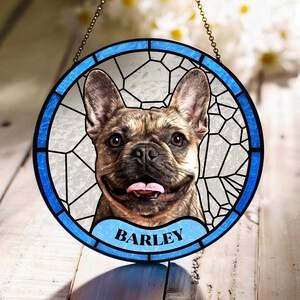 May include: Round stained glass window featuring a smiling brindle French Bulldog with the name "BARLEY" on a blue banner. The window has a blue border and a chain for hanging. The background is a light color.