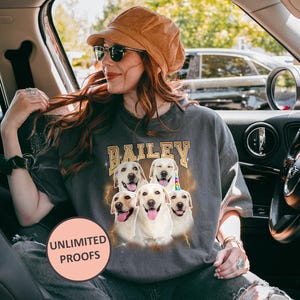 A personalized Comfort Colors 1717 t-shirt in a vintage, faded black or "Pepper" color, featuring a custom digital portrait of a dog on the front, centered on the chest. The dog is a golden retriever with a happy, smiling expression, its fur rendered in warm, natural tones with subtle artistic effects to give it a "bootleg" or retro graphic look. Below the dog's portrait, its name, "Buddy," is written in a distressed, blocky font, consistent with a vintage aesthetic.