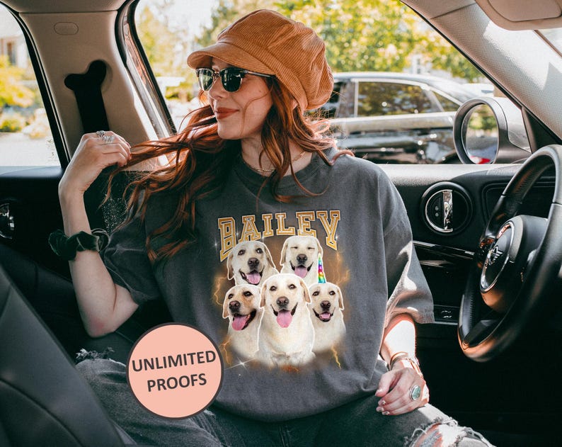 A personalized Comfort Colors 1717 t-shirt in a vintage, faded black or "Pepper" color, featuring a custom digital portrait of a dog on the front, centered on the chest. The dog is a golden retriever with a happy, smiling expression, its fur rendered in warm, natural tones with subtle artistic effects to give it a "bootleg" or retro graphic look. Below the dog's portrait, its name, "Buddy," is written in a distressed, blocky font, consistent with a vintage aesthetic.