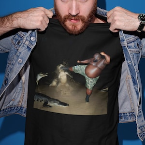 May include: A black t-shirt with a graphic print of a man kicking a crocodile. The man is wearing camouflage shorts and is in a fighting stance. The crocodile is in the water and has its mouth open.