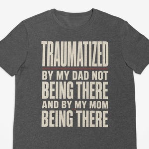 May include: A dark heather gray short-sleeve t-shirt featuring the beige text: TRAUMATIZED BY MY DAD NOT BEING THERE AND BY MY MOM BEING THERE. The shirt has a crew neckline and a standard fit, with a thin horizontal red line below the first word.