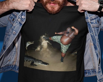 May include: A black t-shirt with a graphic print of a man kicking a crocodile. The man is wearing camouflage shorts and is in a fighting stance. The crocodile is in the water and has its mouth open.