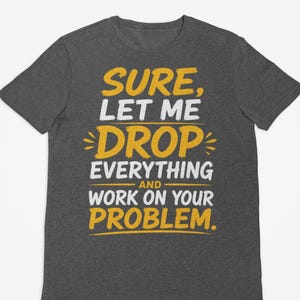 Puede incluir: Una camiseta de manga corta en color gris jaspeado oscuro con el texto en amarillo y blanco: SURE, LET ME DROP EVERYTHING AND WORK ON YOUR PROBLEM. La prenda tiene cuello redondo y corte estándar, con la etiqueta Dark Heather visible.