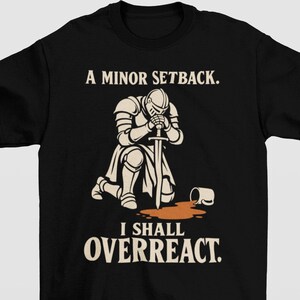 May include: A black t-shirt featuring a graphic of a knight in armor kneeling with a sword, next to a spilled mug of brown liquid. The text reads A MINOR SETBACK. I SHALL OVERREACT. The design is printed in cream and brown tones on the front.