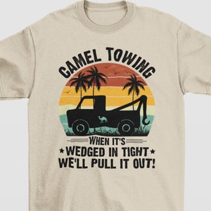 May include: A tan t-shirt with a black graphic design. The design features a tow truck with a camel in the bed of the truck. The tow truck is driving on a beach with palm trees and a sunset in the background. The text on the graphic reads 'Camel Towing When It's Wedged In Tight We'll Pull It Out!'.