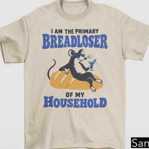 May include: A sand-colored t-shirt featuring a graphic of a black and white mouse wearing blue sunglasses, lounging on a loaf of bread while holding a cocktail. The text reads: I AM THE PRIMARY BREADLOSER OF MY HOUSEHOLD in blue lettering.