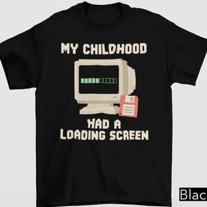 May include: A black short-sleeved t-shirt featuring a pixelated graphic of a beige vintage computer monitor with a green loading bar and a red floppy disk. The text reads MY CHILDHOOD HAD A LOADING SCREEN in a white pixelated font style.