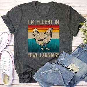 May include: A dark heather grey t-shirt with a vintage-style graphic of a chicken and the text "I'm fluent in fowl language".