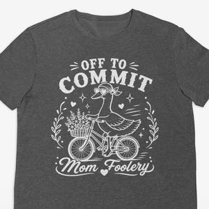 Può includere: Una t-shirt a maniche corte grigio scuro mélange con una grafica bianca di un'oca che indossa occhiali da sole e una bandana mentre va in bicicletta con un cestino di fiori. Il testo recita OFF TO COMMIT Mom Foolery con piccoli cuori.