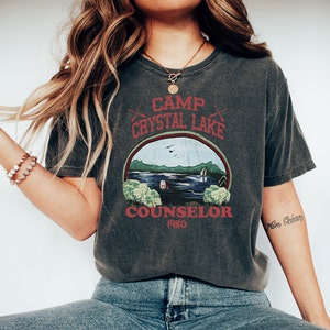 May include: Dark gray t-shirt with a graphic design featuring the text "Camp Crystal Lake Counselor 1980". The design includes a lake scene with trees and a cabin. The shirt has a vintage, worn look.