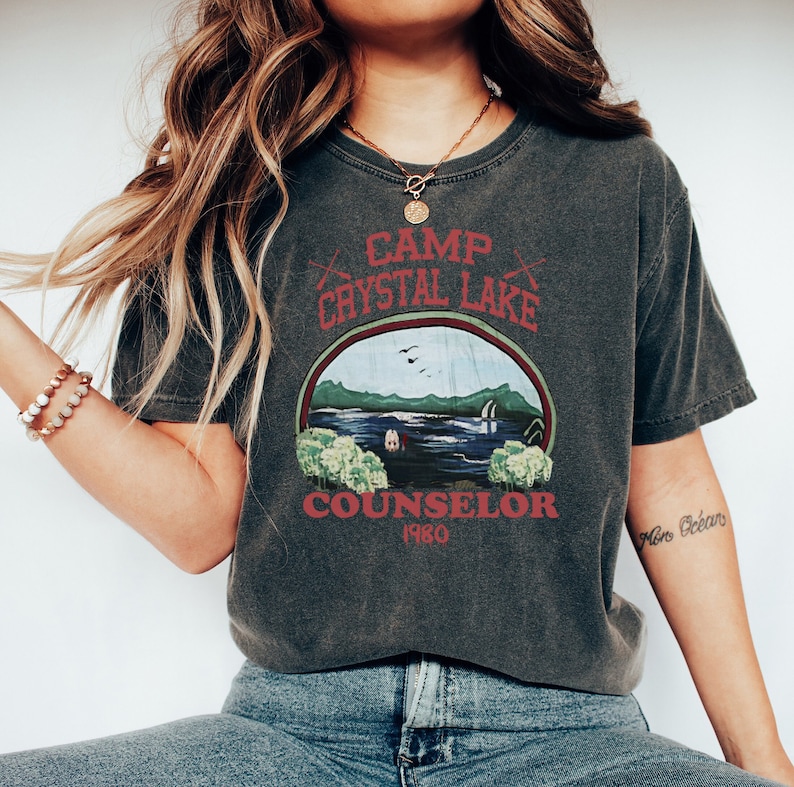 May include: Dark gray t-shirt with a graphic design featuring the text "Camp Crystal Lake Counselor 1980". The design includes a lake scene with trees and a cabin. The shirt has a vintage, worn look.