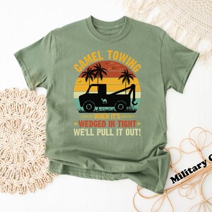 May include: A green t-shirt with a vintage-style graphic of a tow truck pulling a camel. The text reads "Camel Towing When It's Wedged In Tight We'll Pull It Out!"