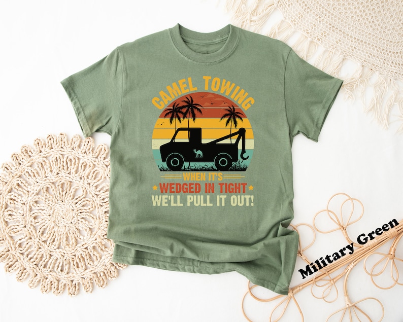 May include: A green t-shirt with a vintage-style graphic of a tow truck pulling a camel. The text reads "Camel Towing When It's Wedged In Tight We'll Pull It Out!"