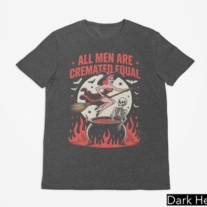 May include: Dark heather gray t-shirt with a Halloween-themed graphic. The design features a witch on a broom, a skeleton, and a cauldron with flames. The text reads "ALL MEN ARE CREMATED EQUAL."