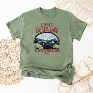 May include: A sage green t-shirt featuring a graphic of a lake scene with the text "Camp Crystal Lake Counselor 1980". The design includes crossed axes and a circular frame around the lake image.