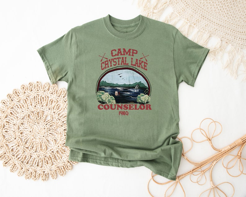 May include: A sage green t-shirt featuring a graphic of a lake scene with the text "Camp Crystal Lake Counselor 1980". The design includes crossed axes and a circular frame around the lake image.