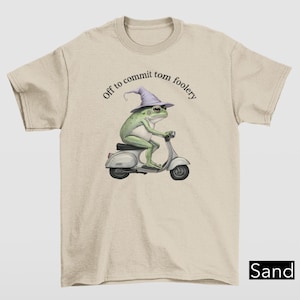 Peut inclure: T-shirt couleur sable avec une grenouille verte portant un chapeau de sorcier violet et conduisant un scooter blanc. Le texte "Off to commit tom foolery" est imprimé au-dessus de la grenouille. Vêtement décontracté.