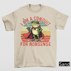 May include: A beige t-shirt with a retro-style graphic of a frog wearing a red cape and holding a sword. The text "I am a conduit for nonsense" is printed below the frog.