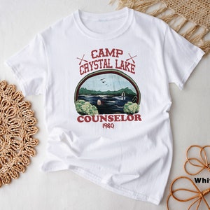 May include: White t-shirt with a graphic featuring the text "Camp Crystal Lake Counselor 1980" in red. The graphic depicts a lake scene with trees, a cabin, and the text "Camp Crystal Lake" in an arch. The shirt is made of a soft material.